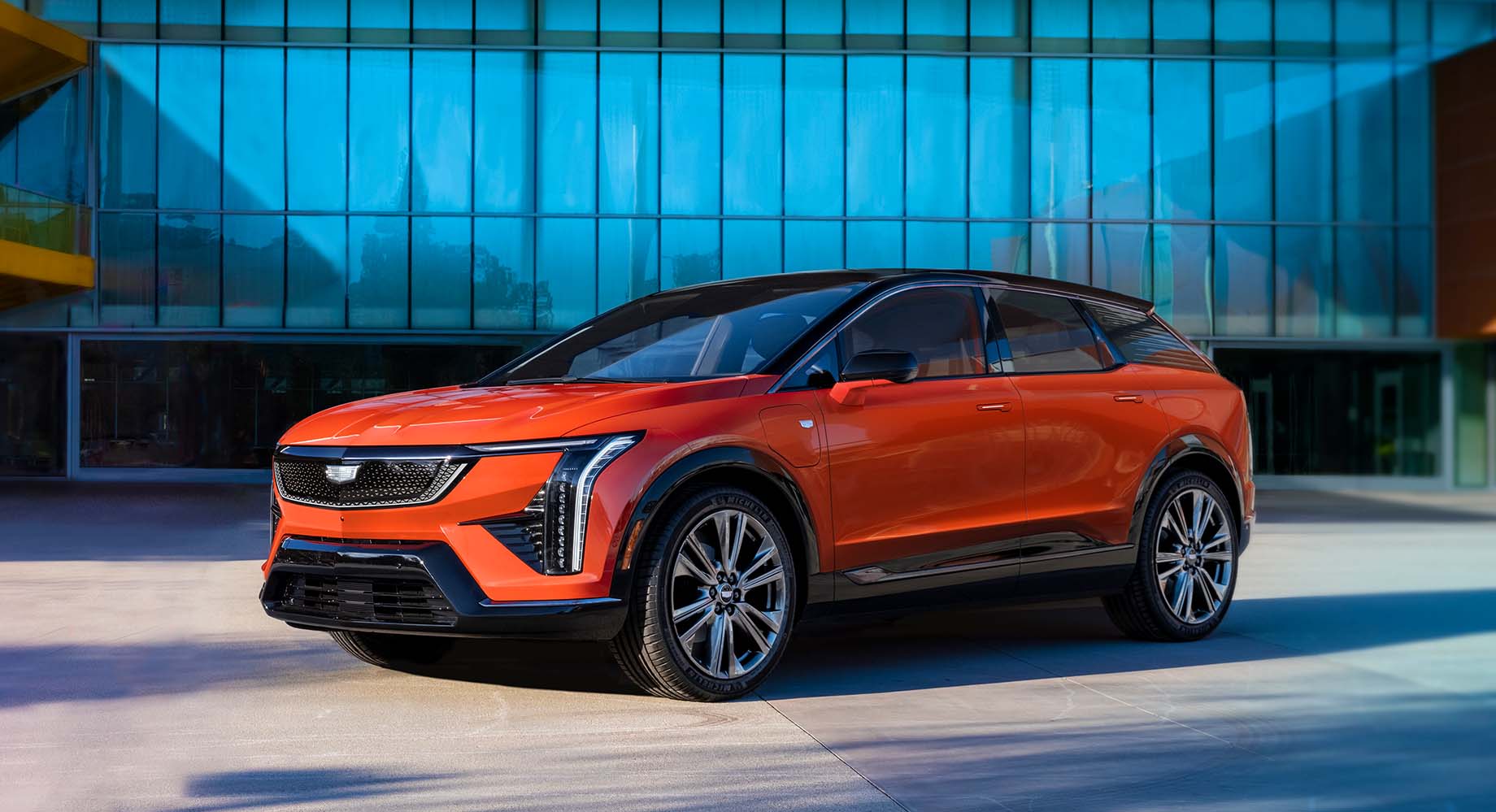 A vibrant orange Cadillac OPTIQ is parked in front of a modern building