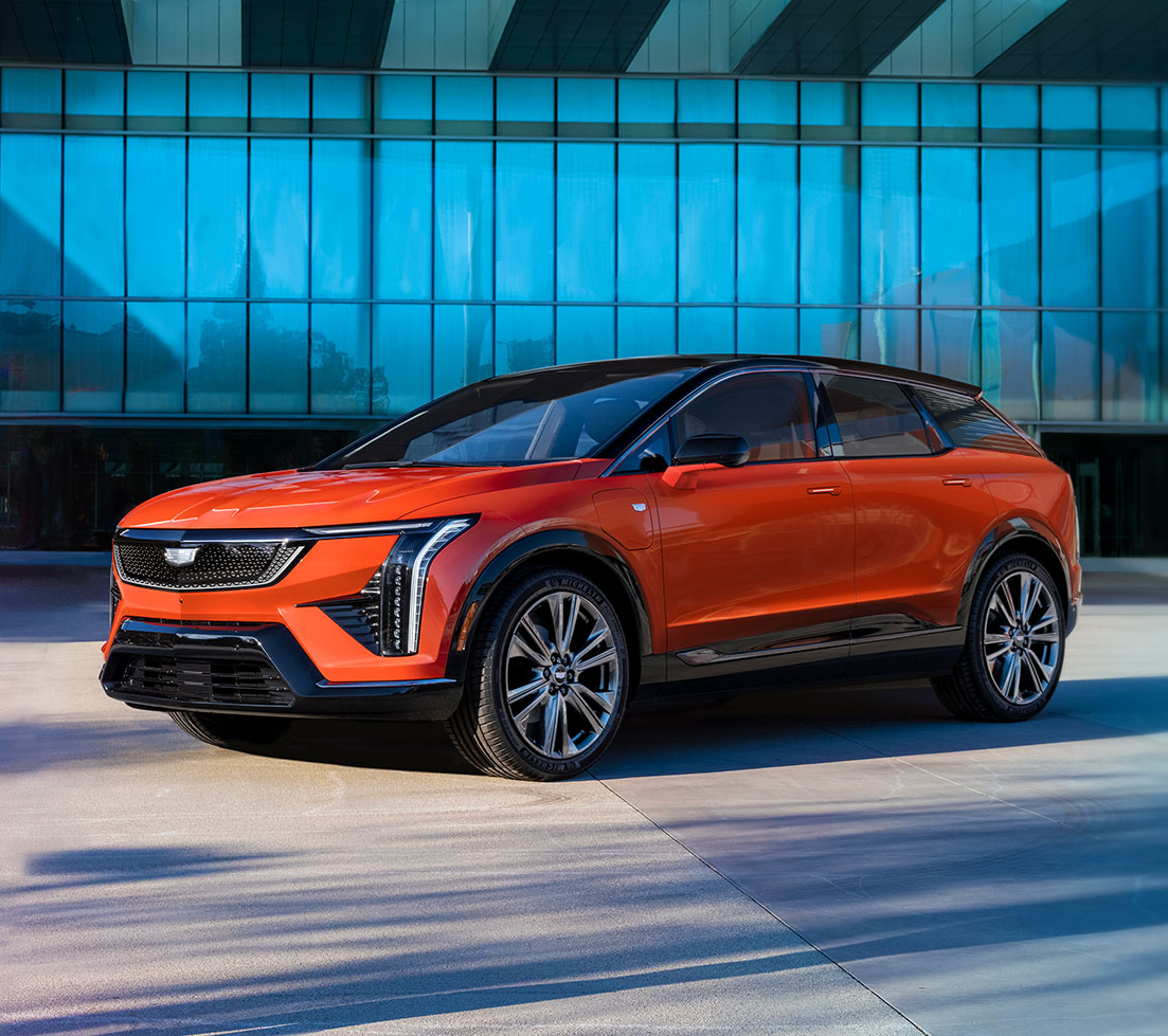 A vibrant orange Cadillac OPTIQ is parked in front of a modern building
