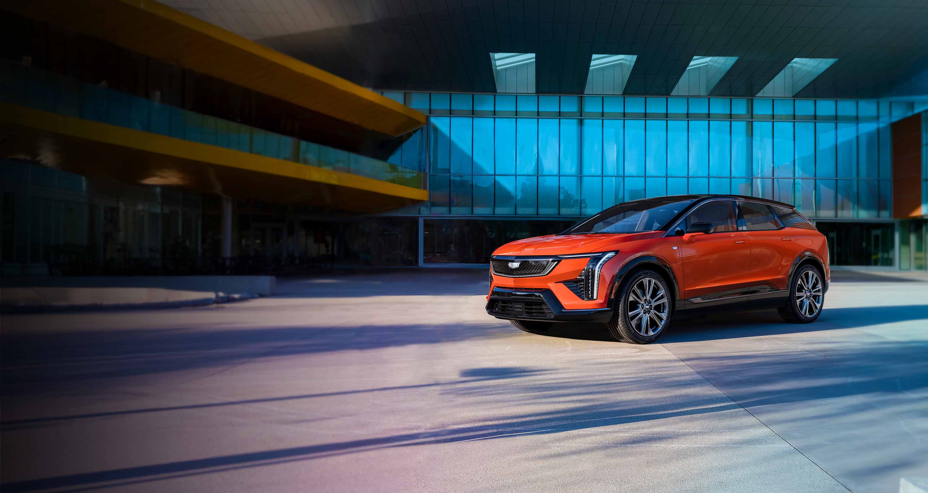 A vibrant orange Cadillac OPTIQ is parked in front of a modern building